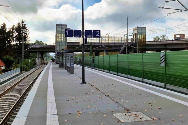Bahnsteige 1 und 2 der Station Frankfurt-Ginnheim mit grüner Lärmschutzwand neben den Gleisen und Fußgängerbrücke und Aufzügen im Hintergrund.