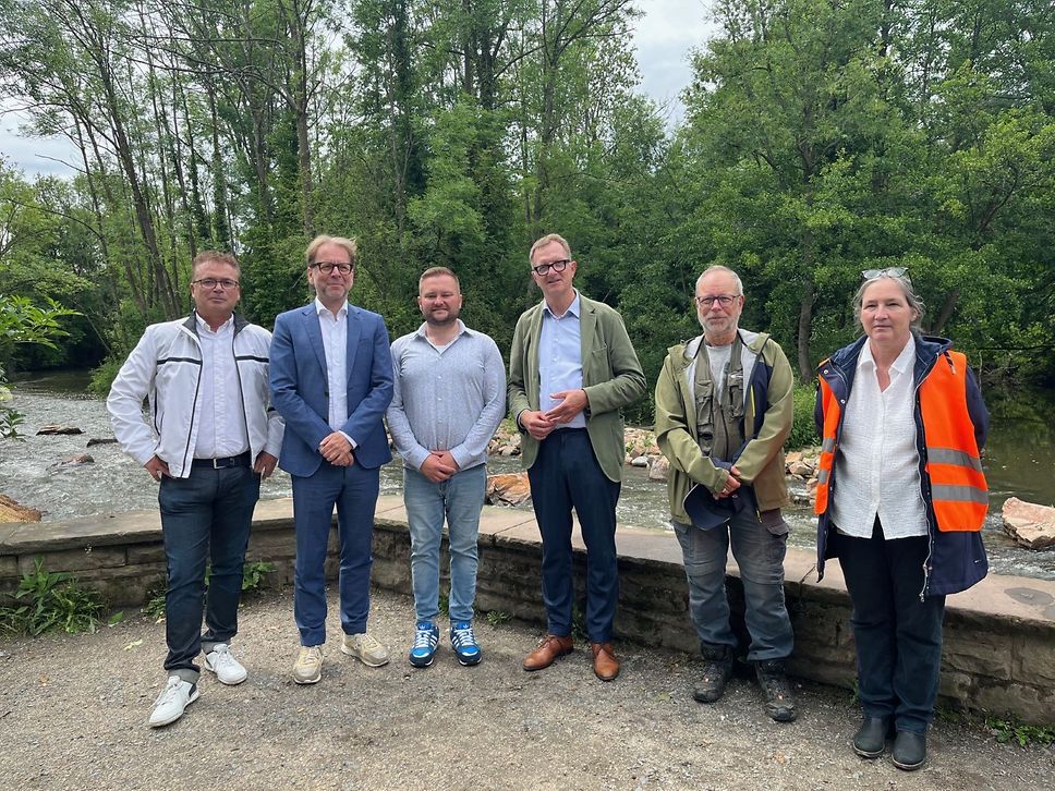 Sechs Personen stehen nebeneinander vor einem Fluss mit Bäumen im Hintergrund. Sie tragen verschiedene Outfits, darunter Anzüge, Freizeitkleidung und eine Warnweste.